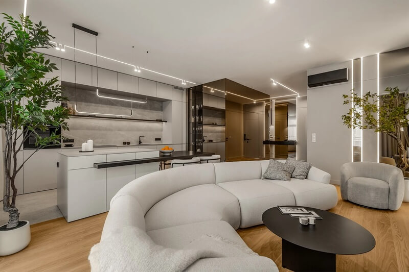 Na zdjęciu widzimy eleganckie, nowoczesne wnętrze salonu połączonego z kuchnią w stylu open space. Dominującym elementem jest duża, jasnoszara sofa o zaokrąglonym kształcie, która tworzy przytulną strefę wypoczynkową. Przed nią znajduje się owalny, czarny stolik kawowy z gazetami i filiżankami. Obok stoi miękki, tapicerowany fotel w podobnym odcieniu co sofa.W tle znajduje się minimalistyczna kuchnia z wyspą i zintegrowanym blatem roboczym, utrzymana w bieli i szarości z elementami czerni. Nowoczesne oświetlenie LED w formie cienkich linii biegnie po suficie i ścianach, nadając wnętrzu futurystyczny charakter. Ciekawym akcentem są wysokie, ozdobne rośliny doniczkowe, które dodają przestrzeni świeżości i naturalnego ciepła.Podłoga z jasnego drewna kontrastuje z nowoczesnymi, surowymi materiałami, tworząc harmonijną i luksusową atmosferę. Całość wnętrza emanuje spokojem, równowagą i elegancją, łącząc nowoczesność z komfortem.
