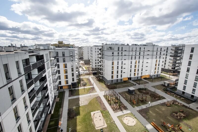 Na zdjęciu widoczna jest nowoczesna zabudowa mieszkaniowa – kompleks wielorodzinnych bloków zlokalizowanych na osiedlu o przemyślanej i uporządkowanej urbanistyce. Budynki mają prostą, geometryczną formę i są utrzymane w jasnej kolorystyce – dominują biel, szarości i elementy grafitu, z akcentami w kolorze żółtym (np. markizy czy detale elewacji).Między budynkami znajduje się przestronny, zadbany dziedziniec z alejkami spacerowymi, placami zabaw, pergolami i terenami zielonymi. Widać ławki, piaskownicę oraz elementy małej architektury. Układ przestrzeni zapewnia mieszkańcom komfort, dostęp do rekreacji i światła dziennego.