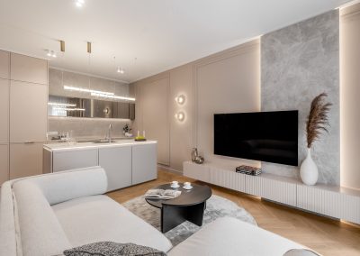 Na zdjęciu widzimy elegancką, otwartą przestrzeń łączącą salon z kuchnią — wszystko utrzymane w stonowanej palecie barw, z przewagą szarości, czerni i bieli. W centrum salonu znajduje się jasnoszara sofa narożna, na której opiera się koc, a przed nią stoi okrągły czarny stolik z filiżankami i magazynem, co nadaje wnętrzu klimat codziennej wygody. Na ścianie zawieszony jest duży telewizor, a pod nim znajduje się biały stolik konsolowy z wazonem i książkami. Część kuchenna prezentuje się równie nowocześnie — wyspa z wbudowanym zlewem i misą owoców stanowi praktyczne i estetyczne centrum. Lustro nad blatem kuchennym odbija wnętrze, dodając przestrzeni głębi, a wiszące lampy nad wyspą stanowią efektowne źródło światła. Całość wyróżnia się przejrzystością, starannie dobranymi detalami i spokojnym klimatem.