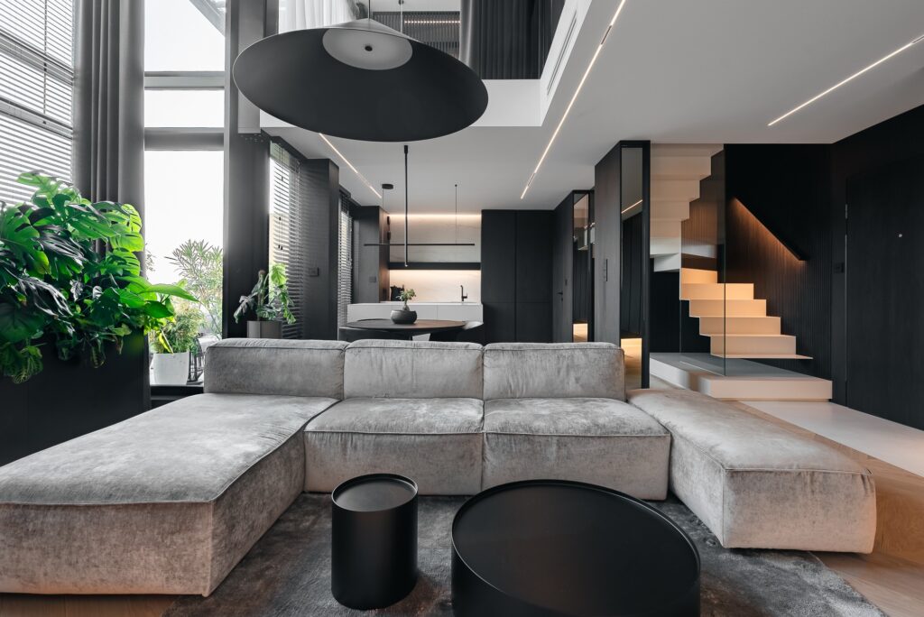 To nowoczesny salon utrzymany w minimalistycznym stylu, w którym dominuje stonowana kolorystyka i eleganckie formy. Centralnym punktem jest duży, szary narożnik z czarnymi stolikami kawowymi, a przestrzeń wypełniają naturalne światło i zielone rośliny. W tle widoczna jest stylowa, czarna kuchnia oraz schody z podświetleniem, prowadzące na górę. Całość sprawia wrażenie przestronności, komfortu i wyrafinowanego designu.